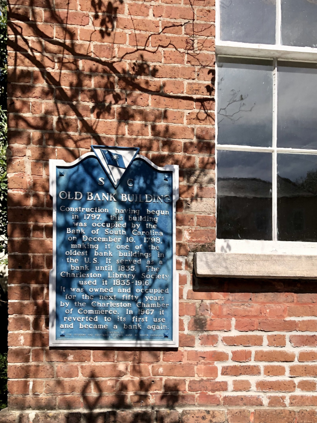 Altes Bankgebäude mit blauer Tafel an der Seite, ein Fenster rechts und ein Baum links; die Tafel enthält Text.