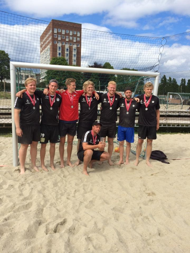 Mannergruppe in Sportkleidung auf einem sandigen Strand, mit Medaillen und einem Netz umgeben, mit Bäumen, Gebäuden und bewölktem Himmel im Hintergrund.