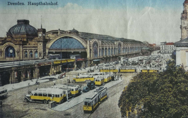 Schwarz-weiß-Postkarte vom Dresdner Hauptbahnhof: Eine belebte Straße mit Fahrzeugen, Fußgängern, Bäumen, Gebäuden und einem bewölkten Himmel.