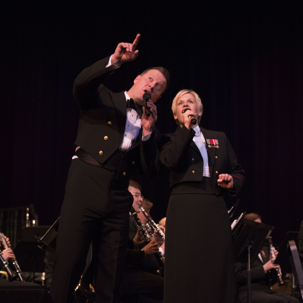 Ein Mann und eine Frau in Smokings mit Mikrofonen auf einer Bühne, mit einem sitzenden Orchester dahinter in einer dunklen Umgebung.