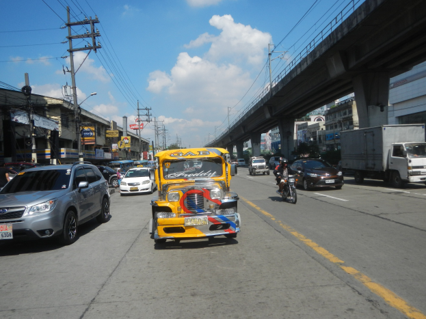 Gelbes Taxi fährt auf einer Straße mit hohen Gebäuden, Strommasten mit Kabeln und einer Brücke im Hintergrund unter einem bewölkten Himmel, mit einem Wasserzeichen auf dem Bild.
