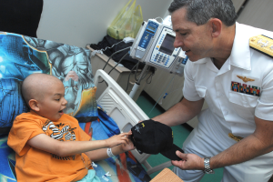 Ein Mann in wei├čer Uniform steht neben einem kleinen Jungen im Krankenhausbett und h├Ąlt eine M├╝tze in der Hand, mit medizinischer Ausr├╝stung und Gegenst├Ąnden im Hintergrund.