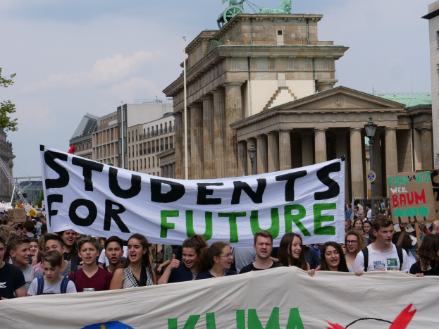 Gruppe von Schülern marschiert in Berlin mit einem bunten "Students for Future"-Schild vor dem Hintergrund von Gebäuden, Bäumen und Himmel.