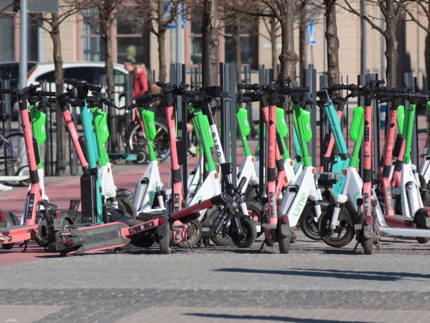 Eine Reihe von Elektro-Rollern, die an einer Straße geparkt sind, mit einer Person im Vordergrund und etwas unscharfen Bäumen und Gebäuden im Hintergrund.