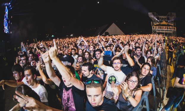 Menschenmenge auf einem Musikfestival mit erhobenen Händen, einige sitzen und andere stehen, in der Nähe eines Metallzauns mit Gebäuden und Lichtern im Hintergrund.