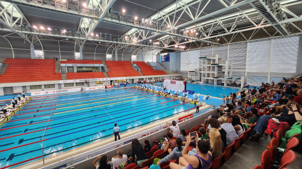 Großer Hallenbad mit vielen Menschen auf Stühlen drumherum und einigen im Wasser Stehenden, mit hoher Decke mit Lampen und Eisenstangen und Schildern im Hintergrund.