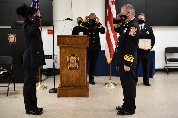 Gruppe uniformierter Personen in Masken auf einem Podium mit einem Mikrofon, umgeben von Fahnen und Möbeln, während einer Vereidigung für einen neuen Feuerchef.