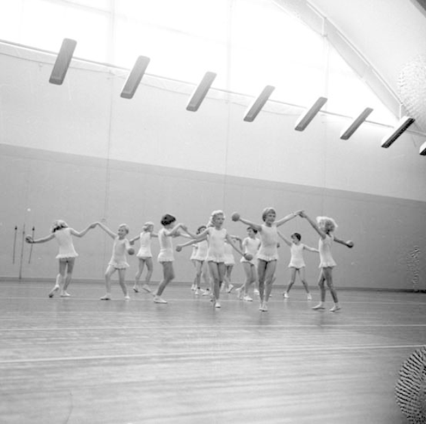Schwarzes Bild von jungen Mädchen in einem Tanzstudio, die sich an den Händen halten und tanzen, mit einem Ball auf der rechten Seite und Lampen an der Decke.
