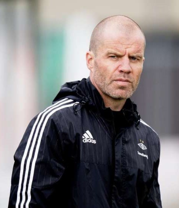 Ein kahlköpfiger Mann in einer schwarzen Jacke steht auf einem Fußballfeld, die Hände vor der Brust verschränkt und einen ernsten Gesichtsausdruck.