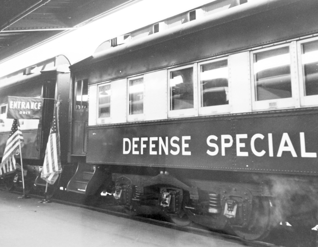 Schwarz-weiß-Foto eines Zuges mit der Aufschrift "Defense Special" auf Gleisen mit zwei Fahnen links und einer Decke oben.