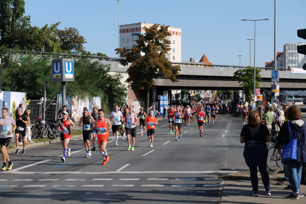 Gruppe von Menschen, die auf einer baumgesäumten Straße mit Laternen, Schildern, Fahrrädern, einem Zaun, Gras, einer Brücke, Gebäuden und einem klaren blauen Himmel laufen.