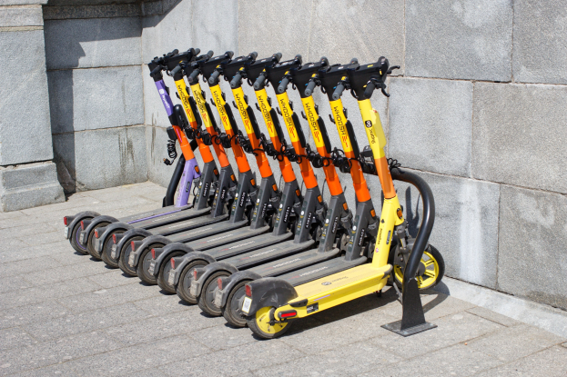 Eine Reihe von Elektro-Scootern, die nebeneinander auf einem Gehweg geparkt sind, mit einer Wand im Hintergrund.