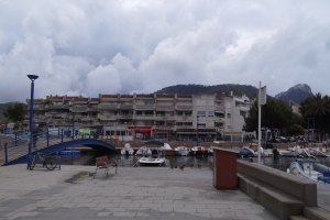 Ein Hafen mit Booten im Wasser, eine Brücke mit Geländern, Bänken, Laternen, Bäumen, Gebäuden mit Fenstern, Hügeln im Hintergrund und einem Himmel mit Wolken.