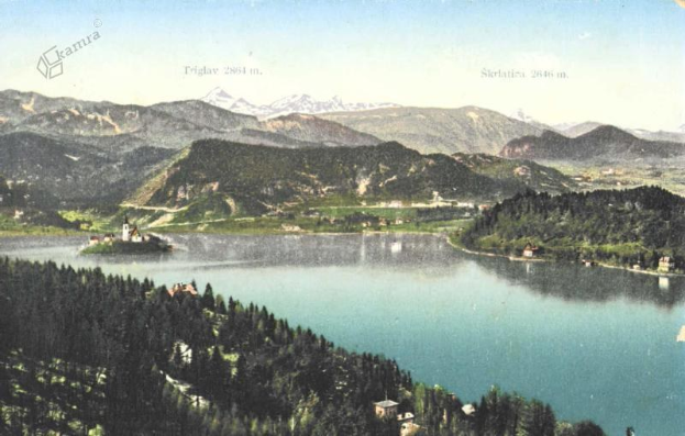 Eine alte Postkarte zeigt einen See umgeben von Bäumen, Häusern und Bergen, mit grünem Gras und weißen Wolken am Himmel und sichtbaren Wasserzeichen.