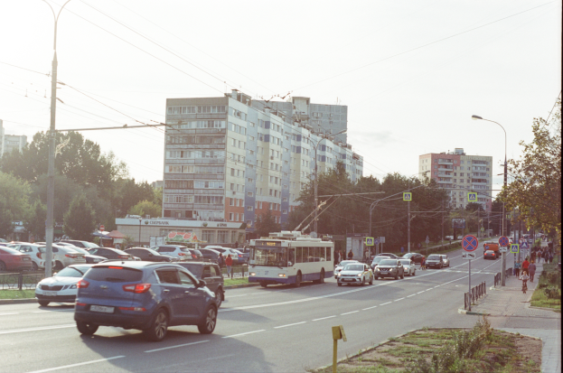 Eine belebte Stadtstraße mit zahlreichen Autos, Bussen, Straßenlaternen, Schildern, Laternenmasten, Strommasten mit Drähten, Bäumen, Gebäuden mit Fenstern unter einem klaren blauen Himmel.