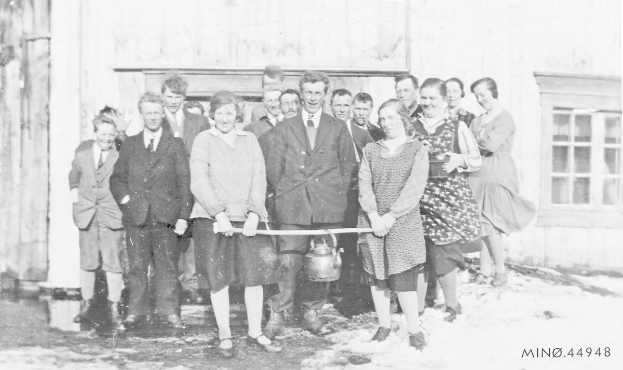 Schwarzes und weißes Foto von Menschen, die ein Band vor einem Schulgebäude mit einem Fenster und einer Tür halten und dabei lächeln.