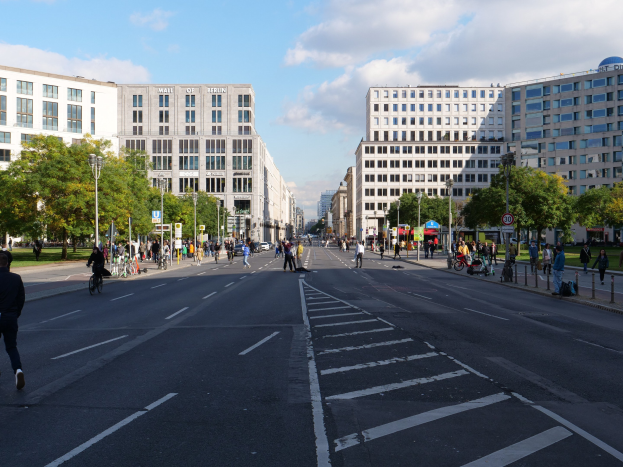 Eine belebte Stadtstraße in Berlin, Deutschland, mit Fußgängern und Radfahrern auf der Straße, umgeben von hohen Gebäuden, Bäumen, Laternen und Schildern unter einem bewölkten Himmel.