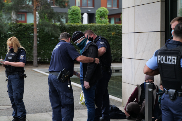 Eine Gruppe von Polizeibeamten vor einem Gebäude stehend, mit einer Frau, die ein Buch und einen Stift hält, und mehreren Personen in der Nähe sitzend und stehend, vor dem Hintergrund von Pflanzen, Bäumen und Gebäuden.