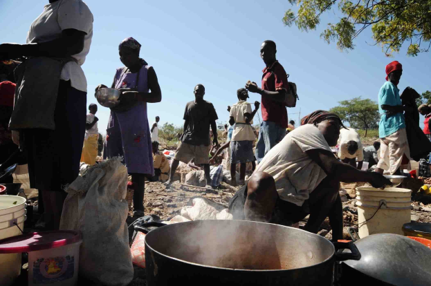 Menschen bereiten Essen im Freien in einer ländlichen Gegend zu, umgeben von Behältern und Materialien, mit Bäumen und einem klaren Himmel im Hintergrund.