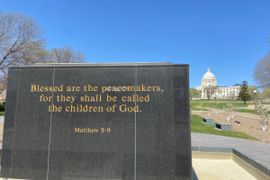 Ein Gedenkstein mit der Inschrift "Selig sind die Friedfertigen, denn sie sollen das Reich Gottes erben" umgeben von Bäumen, Pflanzen, Gras, Laternenmasten und einem Gebäude unter einem klaren blauen Himmel.