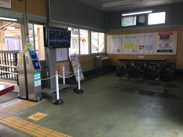 Ein belebter Bahnhofseingang mit Reihen von Stühlen, einem Service-Schalter, Ticketautomaten, Kettenpfählen, Informationsanzeigen, Wandplakaten, Deckenbeleuchtung und Geländern.