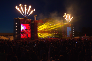 Eine große Menschenmenge versammelte sich auf einem Musikfestival, mit einer Bühne in der Mitte, auf der Musiker Instrumente spielten, beleuchtet von hellen Lichtern und umgeben von Bannern, Zelten links und einem dunklen Himmel mit Feuerwerk im Hintergrund.