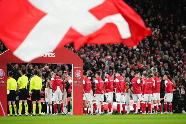 Gruppe von Menschen auf einem Fußballfeld mit einer roten und weißen Flagge im Vordergrund, einem Bogen mit der Aufschrift "Bayern München vs. Bayern München Betting Tips & Preview" im Hintergrund und einer großen Stadion-Menge.