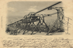 Ein altes Schwarz-Weiß-Foto einer Eisenbahnbrücke mit einem Kran, umgeben von Bäumen und einem bewölkten Himmel, mit Text unten, der besagt: "Bahnstrecka, die erste Eisenbahnbrücke in Deutschland".