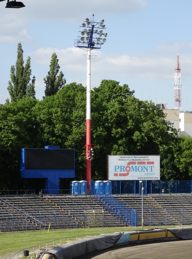 Rennstrecke beleuchtet von oben, umgeben von Gras, Geländern, Treppen, einem Lichtmast, einem Schild, Bäumen, Gebäuden, Türmen und einem bewölkten Himmel.