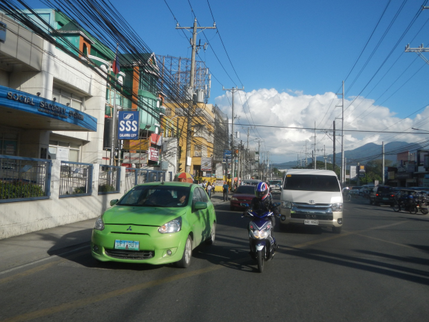 Grünes Auto fährt eine Straße mit hohen Gebäuden, Strommasten, Fußgängern, Bäumen, Bergen und Wolken im Hintergrund.