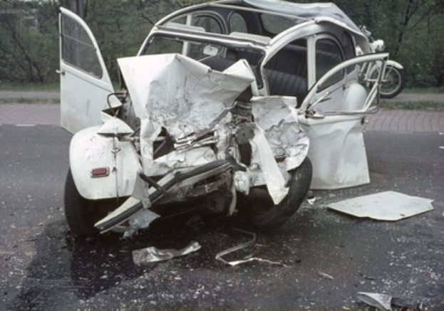 Ein verunfalltes Auto mit schwerer Frontschaden steht am Straßenrand, mit Bäumen, einem Gebäude und einem weiteren Fahrzeug im Hintergrund.