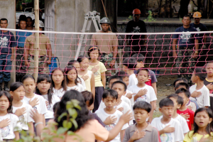 Gruppe von Kindern vor einem Netz mit einer Frau im Vordergrund, Menschen mit Helmen, einer Leiter, einer Wand und Bäumen im Hintergrund.