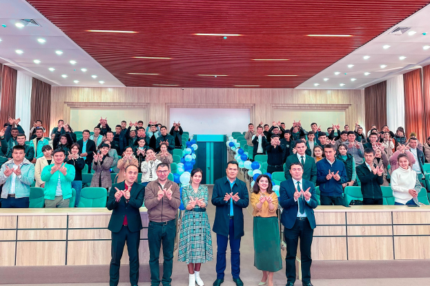 Eine Gruppe von Menschen steht vor einer Menge, einige klatschen, mit Stühlen, Tischen, Ballons, Vorhängen, einer Wand im Hintergrund und Lampen an der Decke, bei der Eröffnungsfeier einer neuen Schule.