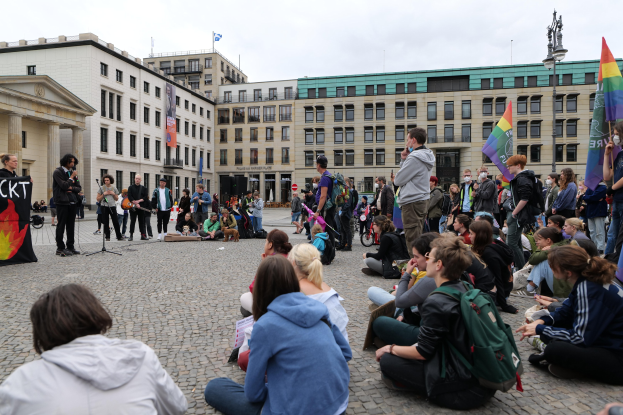 Eine Gruppe von Menschen sitzt auf dem Boden vor einer Menge, die Fahnen und Transparente schwingt, bei einer Demonstration in Berlin mit einer Statue, Gebäuden und einem bewölktem Himmel im Hintergrund.
