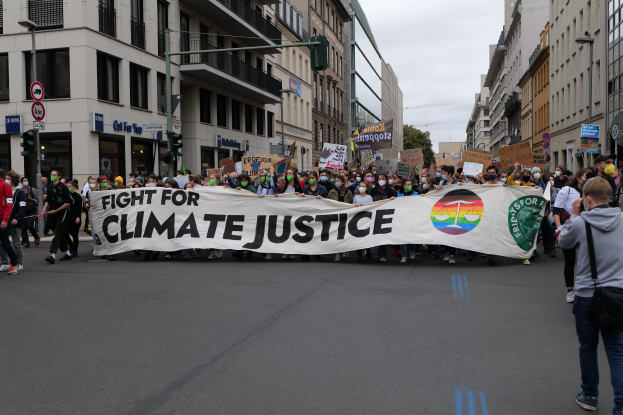 Eine Gruppe von Menschen marschiert mit einem "Kampf für Klimagerechtigkeit"-Schild die Straße entlang, mit Gebäuden, Schildern, Verkehrsampeln und Bäumen an der Straße unter einem bewölkten Himmel.