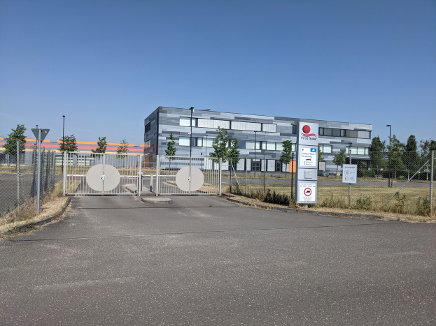 Großes Industriegebäude mit der Aufschrift "First Solar" und vielen Fenstern, umgeben von einem Metallzaun, Bäumen, Pflanzen, Gras, Schildern, Straßenlaternen und einem Weg unter einem bewölkten Himmel.