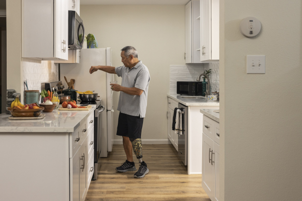 Ein Mann steht in einer Küche mit weißen Schränken und hält ein Glas in der Hand, während verschiedene Gegenstände, einschließlich Obst und Küchenutensilien, auf einer Arbeitsplatte zu seiner Linken liegen.