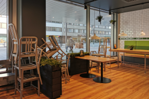 Ein Restaurant mit Holztischen und -stühlen vor einem großen Fenster, umgeben von Pflanzen und Lichtern, mit Menschen und Gebäuden draußen in Berlin, Deutschland.