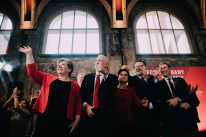 Eine Gruppe von Menschen steht vor einer Menge und klatscht feierlich, mit einem Banner, Glasfenstern und beleuchteten Lichtern im Hintergrund, was auf eine Labour Party-Siegfeier für die Wahl 2019 hinweist.