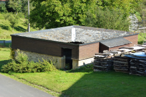 Kleines hölzernes Gebäude neben einer Straße, umgeben von Gras, Pflanzen, Bäumen und einem Pfahl, mit gestapelten Holzscheiten auf der rechten Seite.