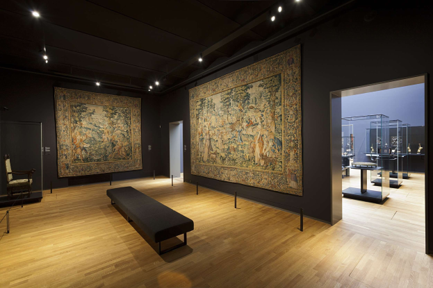 Eine Galerie im Museum of Fine Arts, New York, mit zahlreichen Gemälden an den Wänden, einer Bank, Pfosten mit Seilen, einem Stuhl, einer Tür, Deckenleuchten und verschiedenen Gegenständen auf den Tischen.