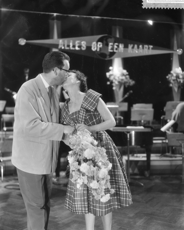 Ein Mann und eine Frau umarmen und küssen sich auf einer Bühne, wobei die Frau einen Blumenstrauß hält und im Hintergrund Stühle und Blumenvasen zu sehen sind.
