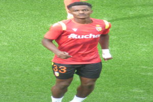 Emmanuel Adebayor, ein Mann in rotem T-Shirt und schwarzen Shorts, geht über ein Fussballfeld, mit zwei Personen im Hintergrund.