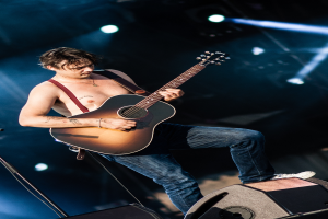Ein Mann ohne Hemd spielt eine Gitarre auf der Bühne bei einem Konzert, beleuchtet von Scheinwerfern im Hintergrund, vor einem Mikrofon mit Ständer und Lautsprechern am Boden.