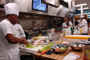 Eine Gruppe von Köchen in einer Küche, die Essen auf einem Schneidbrett zubereiten, mit einem Koch im Vordergrund in einer weißen Schürze und Mütze, der ein Messer hält, umgeben von Utensilien, Schüsseln, Gemüse, einem Handy, Flaschen und anderen Gegenständen, mit einem Fernseher und Deckenleuchten im Hintergrund.