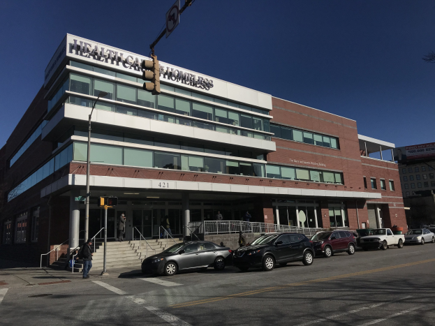 Großes Gebäude mit der Aufschrift "New York City Health Care Homeless Center" mit parkenden Autos davor, umgeben von Straßeninfrastruktur und Passanten unter einem klaren blauen Himmel.