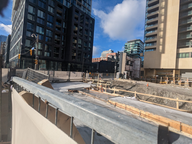 Baustelle in Toronto mit einem großen Gebäude im Bau, umgeben von Absperrungen, Pfählen, Verkehrszeichen und Schildern, unter einem bewölkten Himmel.