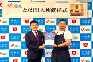 Zwei lachende Männer mit einem Zertifikat, im Hintergrund ein Banner mit der Aufschrift "Toda City Wins PR Award" und ein Foto einer Person.