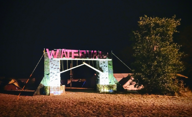 Bühne für ein nächtliches Musikfestival mit einem Bogen mit Text, einem Baum auf der rechten Seite, Zelten im Hintergrund und einem dunklen Himmel.