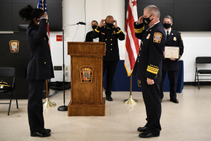Gruppe uniformierter Personen in Masken vor einem Podium mit Mikrofon, umgeben von Flaggen und Möbeln, während einer Amtseinführung, bei der eine Person ein Zertifikat hält.